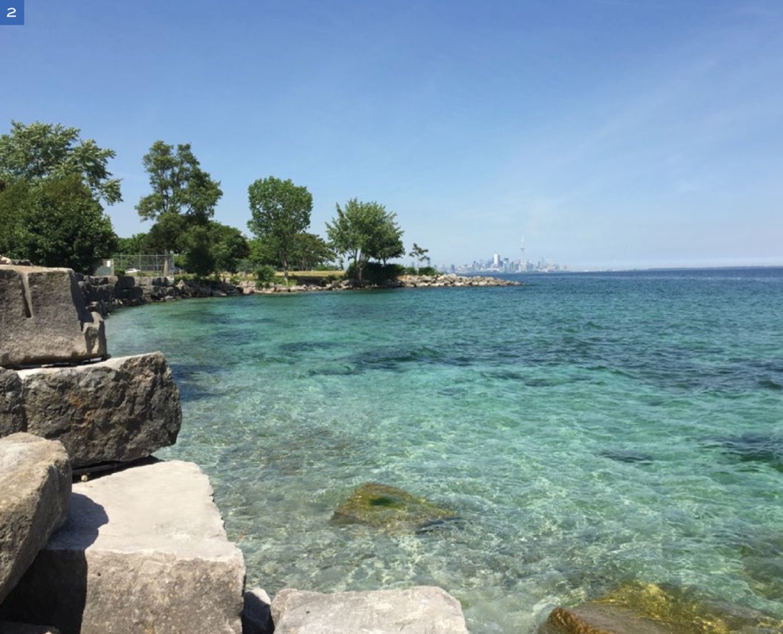 Clear turquoise water along the rocky Long Branch shoreline with the Toronto skyline in the distance